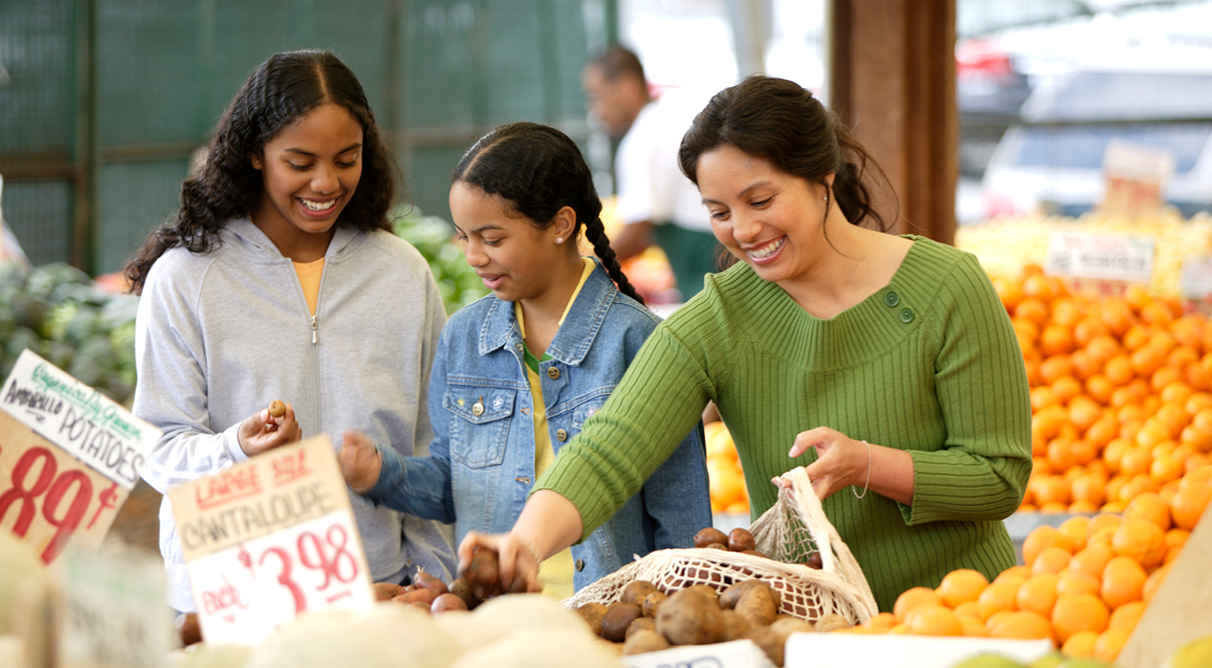 Una mujer sonríe y dos niñas jóvenes compran en el puesto de frutas y verduras.